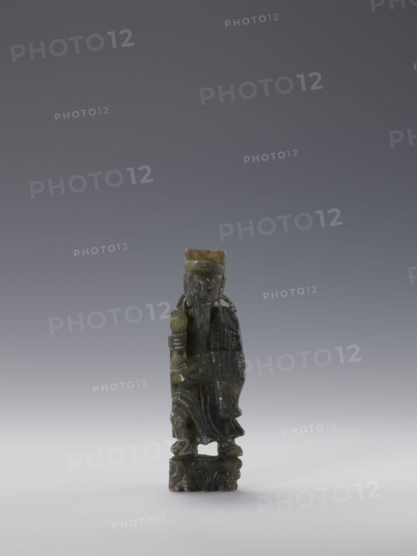 Sage, unknown sculptor, figurine, Chinese culture, Chinese art, sages, Qing dynasty 1644–1911, grey-green soapstone, 19th century, China, sculpture; height 13.8 cm, width 4.4 cm, depth 3.1 cm, A small carved figurine stands on a carved base, the figure wearing a long flowing robe and a tall hat, holding a rounded object in one hand and a staff or similar object in the other; the carving shows facial features and a long beard, with draped sleeves and detailed folds in the clothing. The object is primarily shades of dark green and olive with some lighter greenish-yellow areas and slight mottling, set against a neutral gradient background of gray transitioning to a pale gray near the bottom.