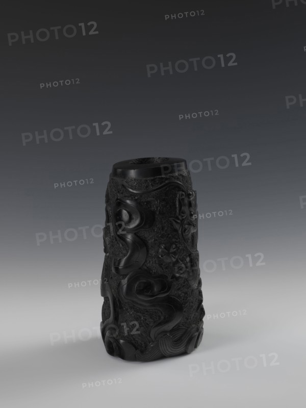 Vase, gift, Chinese art; Chinese; Qing dynasty 1644–1911; 19th century. Vessel with bas-relief decoration of dragons. Height 24.5 cm; diameter 14.3 cm., A single cylindrical carved object stands upright on a smooth surface, appearing dark black with a glossy sheen and matte textured areas; its surface is covered in raised swirling and organic relief patterns that form flowing curves and floral motifs, with areas of rough stippled texture contrasting with the polished carved sections; the top rim is visible and flat, and the base sits casting a soft shadow; the background shows a gradient from dark gray at the top to lighter gray near the bottom.