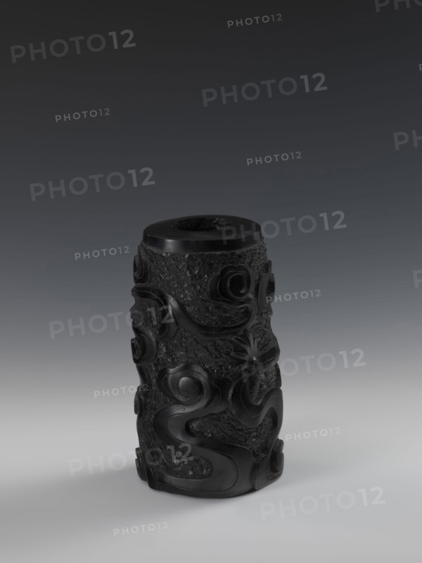 Vase, Chinese, Qing dynasty 1644–1911, 19th century. Bas-relief decoration with dragons iconography. Vessel, height 24.5 cm, diameter 14.3 cm., A dark cylindrical carved object stands upright on a smooth surface, showing a textured body with raised swirling and curving motifs that wrap around it; the material appears glossy in places and more matte and rough in others, with visible variations in surface texture. The top has a recessed circular opening and a smoother rim. The lighting casts soft shadows beneath the object. The visible colors in the image are black and shades of gray.