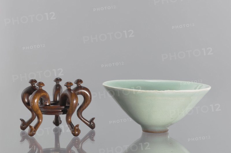 Bowl on base, maker, Shallow, round, profiled base with a round hole in the center; mounted on five legs formed as stylized geese with bent necks protruding above the base. Chinese celadon ceramic vessel, gray-green glaze; Qing dynasty 1644–1911, 18th–19th century. Height 6.1 cm; diameter 12 cm., A small wooden stand with a warm brown finish and curved, ornate legs sits next to a pale green glazed ceramic bowl; the stand has a glossy surface and rounded top elements, and the bowl has a smooth, slightly flared rim and a soft, matte-to-gloss gradient on its exterior, with both objects reflected on a shiny gray surface and set against a neutral light gray background, showing colors including brown, pale green, gray, and hints of white.