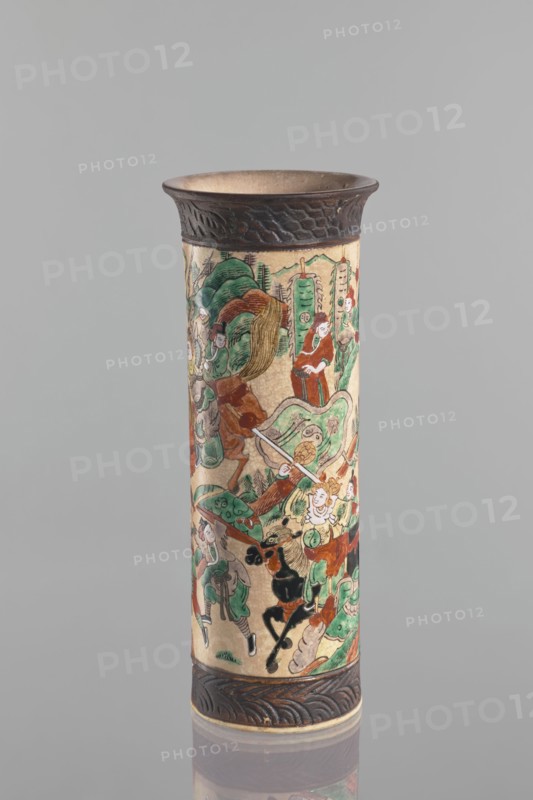 Vase, Chang author's role classification, workshop author; knightly scenes; ceramics; Chinese art; Chinese culture; cylindrical vessel with flared rim; cracked iron-sand glaze; decorated with a scene of a knightly procession in a landscape a mirror equivalent of the decoration on the second vase in the pair; band imitating a bronze representation at the foot and rim; 19th century, China; vessel; height 31 cm, diameter 12 cm., A tall cylindrical object with decorative bands at the top and bottom in dark brown, featuring a painted scene around its body in colors including green, red, brown, black, ivory, and touches of pale yellow; the painted area shows a group of human figures and animals with stylized foliage and abstract shapes, some figures wearing robes and holding long objects, with flowing lines and curved forms; the top band has a textured, carved pattern and the bottom band mirrors this carved motif; the background behind the object is a smooth light gray.