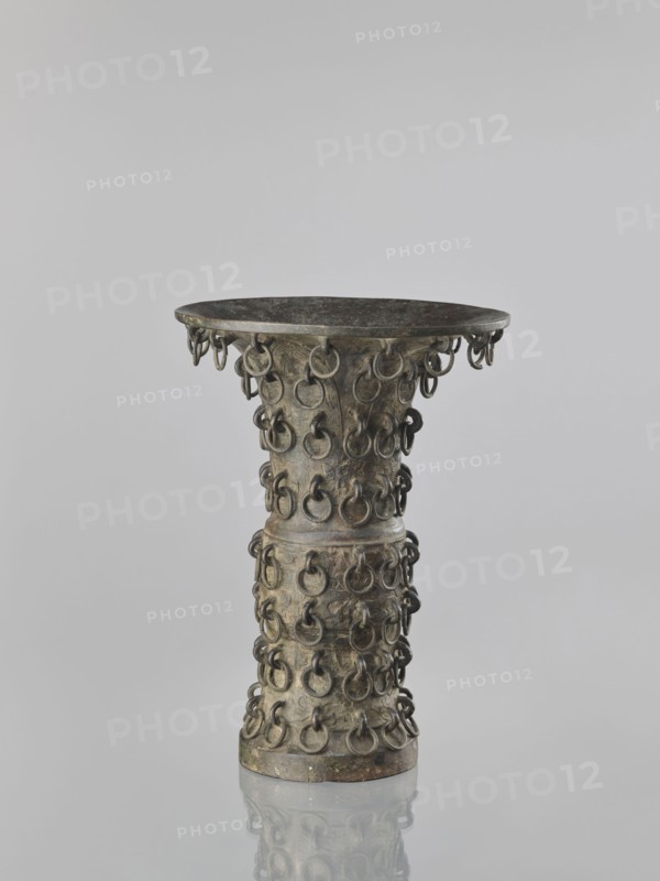 Zun-type vase, Chinese, 19th century Qing 1644–1911, ceremonial/ritual vessel, taotie iconography, vessel, height 33.0 cm, diameter 23.8 cm, A tall, hourglass-shaped metal vessel with a wide, shallow rim and a narrow waist is shown standing on a reflective surface; its surface is studded all around with a repeating pattern of small metal rings attached in rows, and the metal shows a patina with visible greenish and brownish tones along with darker gray and black areas.