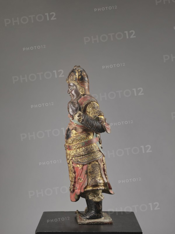 Lokapala — Guardian of the World; creator, Chinese Buddhism; figurine sculpture of a warrior in polychrome red, brown, black; Qing dynasty 1644–1911; 19th century ; full sculpture; height 72.5 cm; China; material: sculpture., A painted and gilded standing figure of a warrior shown in profile, wearing an ornate helmet and layered armor with intricate decorative patterns; the figure has a contemplative facial expression with arms positioned in front, and a draped skirt-like lower garment with visible folds. The visible colors include gold, red, black, brown, and traces of green, with worn areas showing a darker underlayer. The figure stands on a small rectangular base placed on a flat black plinth against a neutral gray background.