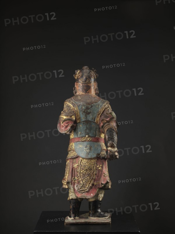 Lokapala Guardian of the World, unknown sculptor; Chinese; Chinese Buddhism; figurine of a warrior; Qing dynasty 1644–1911; polychrome full sculpture in red, brown, and black; 19th century ; China; height 72.5 cm., A painted statue viewed from behind showing a robed figure standing on a rectangular base against a dark background, with visible colors including blue, red, gold, black, green, and brown; the figure wears layered garments with ornate decorative patterns and raised relief details, a belted waist, a lower robe with textured scale-like motifs, and sleeves with folds, and has a helmet or headdress with sculpted elements and worn paint; the boots are black and the base shows scuffed paint.