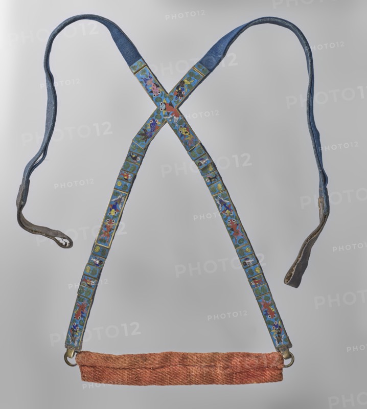 Tailpiece, repossession provenance; Chinese; Qing dynasty 1644–1911; riding equipment for horses; cloisonné enamel; fish motif; made of leather ribbon and bronze decorated with cloisonné enamel on copper colorful, blue background; 18th century; China; horse tack element; length 110 cm, A pair of crossed straps forming an X shape with attached hanging ends and a horizontal padded section at the bottom; the straps are primarily blue with decorative rectangular panels featuring colorful floral and animal-like motifs in shades of blue, green, red, yellow, white, and black, and edged with thin metallic trim; the hanging ends show worn darker material and metal fittings; the bottom padded section is a textured reddish-orange fabric; the background is a smooth light gray.