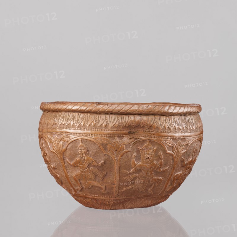 Bowl, 19th century, India. Vessel height 11.5 cm, diameter 18.1 cm. Depicts figures of the god Vishnu placed in arcade poses; Vishnu avatars, turtles; lions, fish, wild boars, flood scenes; trees, plant ornaments, geometric ornaments. At the top and bottom an ornament of stylized palm leaves. Makers unknown., A carved bowl with a rounded shape in a brownish-copper color sits against a plain light gray background; the bowl has a textured rim with a twisted rope-like pattern and a band of carved leaf-like designs below it, while the body features a repeating series of arched panels each containing a carved human figure in dynamic poses, with additional carved foliage and decorative borders around the base, all in similar brown and copper tones.