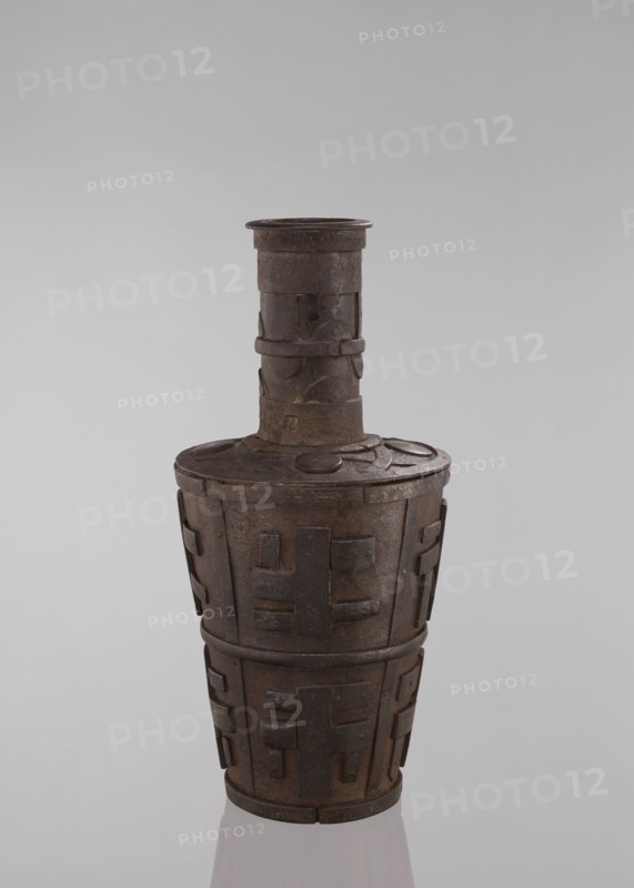 Jug, vessel tapering towards the bottom, Tibetan, 18th–19th century, ceramic vessel, height 46.3 cm; features meander ornament, symbolic motifs, Buddhist art, vases., A tall, narrow vessel with a long cylindrical neck and a wider, tapered body sits against a plain light gray background. The surface of the object appears textured and metallic with visible wear and a patina of dark brown and rust tones, along with areas of darker gray. The neck has a few raised bands and rectangular applied elements, while the shoulder and body display repeated raised geometric motifs and rectangular panels in the same dark brown and gray palette. The top rim is slightly flared and shows the same worn metallic coloration, and the base sits on a reflective surface that picks up faint gray reflections.
