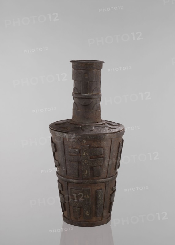 Jug, vessels, Tibetan, vases, meander ornament, symbols, Buddhist art. Vessel tapering towards the bottom. 18th–19th century, Tibet, vessel. Height 46.3 cm., The image shows a tall, narrow vessel with a long cylindrical neck and a wider, rounded body, rendered in dark brown metal with areas of lighter brown and subtle greenish patina; the surface features raised geometric and rectangular relief panels and bands that wrap around the body and neck, and a few circular disc motifs on the lid area; the vessel rests on a reflective pale surface against a smooth light gray background.