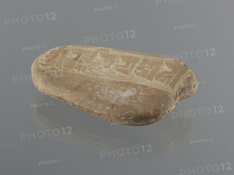 Mani stone, Tibetan Buddhist art, Tibetan culture, Buddhism, mantras, inscriptions, ritual stone, 19th/20th century, Tibet, cult object; weight 1.04 kg, height 16.7 cm, width 10.5 cm, A smooth, rounded flat stone rests on a reflective gray surface, showing a worn top with lighter scratches and incised patterns. The stone is mostly tan and brown with areas of pale beige and faint white scuffing. Its edges are slightly darker brown and one end shows a small rounded notch with darker brown discoloration. The surface reflects softly below, producing a muted mirrored shape in shades of gray and brown. The background is a uniform mid-gray.