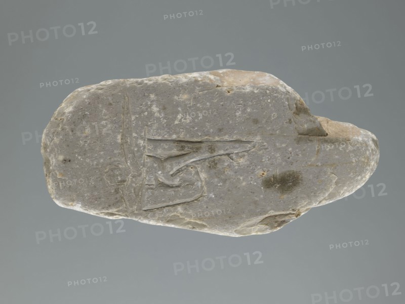 Mani stone, gift; Tibetan Buddhist mani stone with inscriptions of mantras; ritual cult object, stone slab; Tibetan, 19th–20th century; material: stone; weight 0.7 kg; height 18.3 cm; width 9.0 cm., A single irregularly shaped stone rests against a plain gray background; the stone has a muted palette of light gray with areas of beige and pale brown, mottled with small lighter flecks and a few darker gray spots. The stone’s surface is uneven with smooth worn areas, shallow grooves, and a few incised linear marks forming angular shapes. Edges of the stone are rounded in places and slightly jagged in others, showing subtle texture variations and faint patches of discoloration in beige and brown tones. The overall appearance is of a weathered, pale gray rock with beige highlights and scattered darker gray blemishes.