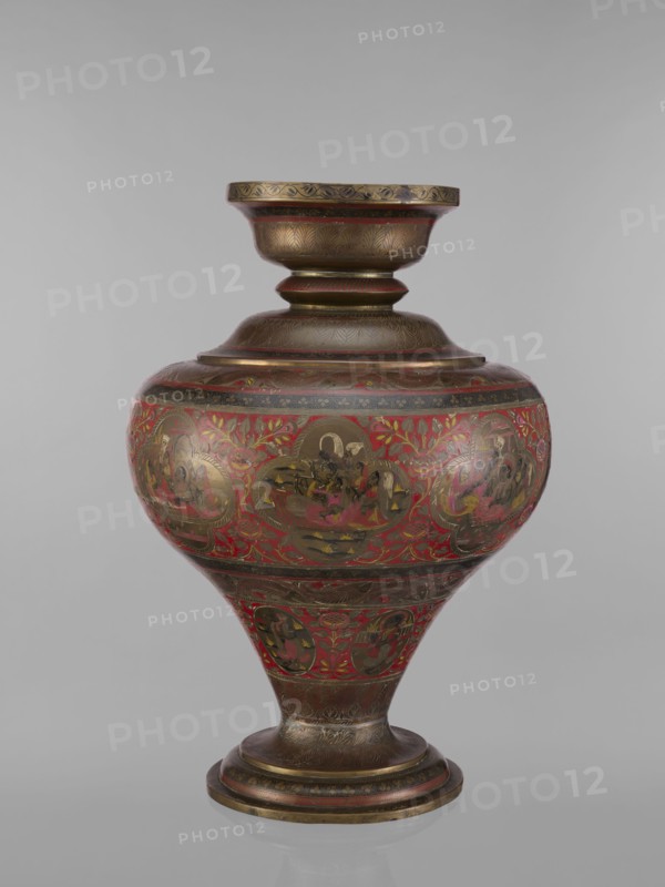 Vase, 19th century, India. Vase with a bulging, flattened belly tapering toward the bottom; wide spout and round foot. Polychrome in red, pink, yellow, white and black. Height 50.0 cm, diameter 13.0 cm, weight 5.75 kg. Decorative program: Hindu subjects Vaishnavism, Shaivism depicting gods Vishnu, Shiva, teachers, ascetics; motifs include fish, plant and geometric ornaments; jugs/containers., An ornate rounded vessel with a flared neck and pedestal base sits against a plain gray background; its surface displays a dominant red color with extensive gold and dark brown metallic detailing, including patterned bands and decorative floral motifs, and several medallion-like scenes rendered in darker tones surrounded by gold highlights; the neck, shoulder, and base show etched or engraved feather-like patterns in brown and gold, while the midsection has red panels filled with stylized leaves and floral elements in gold and darker red, and the foot and lip carry concentric bands of brown and go...