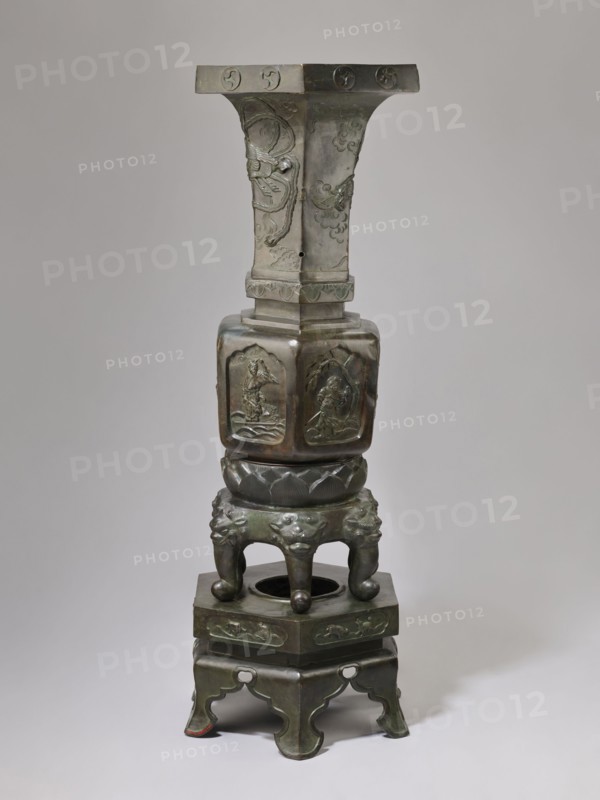 Vase-shaped stove, Chinese art, Chinese culture, unknown provenance; vase-shaped stove decorated with plant ornament, pines, phoenixes, tigers, lotus throne, gourds, dragons, sages, vipers, warriors, lions; associated with Chinese Buddhism, Taoism, and Immortals iconography; Qing dynasty 1644–1911, 19th century, China; height 138 cm, width 42 cm, weight 65.5 kg., A tall sculptural object made of dark greenish bronze with areas of brownish patina stands against a neutral light gray background; its form includes a wide flat top with carved circular motifs, a flared central shaft with raised decorative reliefs and engraved cloudlike patterns, a hexagonal midsection with carved panels depicting human figures in relief, a rounded lotus-shaped collar, and a base formed by a group of curved legs with lionlike faces and ball-shaped feet resting on a stepped plinth that has carved decorative bands and scalloped cutouts.