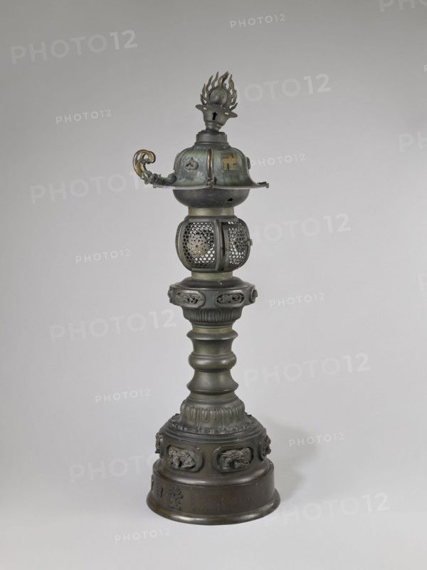 Temple lantern, Japanese, Edo period Man'en, 1860–1861; 1860, 9th month of the Year of the Monkey. Bronze; lantern on a high, multi-segmented shaft with a large round base; hexagonal roof decorated with dragons, pendants and bells at the eaves, topped with a flaming pearl. Assembled from elements cast in bronze, with relief and applied solid decorations. Iconography: dragons, swastika, monkey zodiac; function: temple lantern. Height 184.0 cm; weight 54.0 kg., A tall metal object stands against a plain light gray background, showing a vertical form made of a dark greenish-bronze metal with areas of darker patina and hints of muted gold. The top section features a flame-shaped finial above a rounded cap with a curved decorative handle on one side that has a more golden color. Below that is a spherical cage-like element with perforated decorative patterns. The central shaft is composed of multiple rounded and fluted segments with a ribbed collar, all in the same dark metallic green and br...