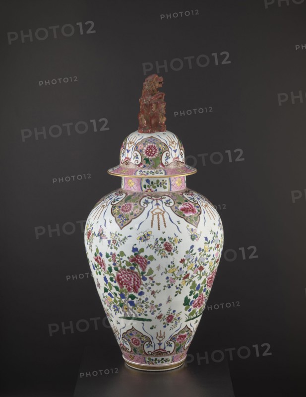 Guan vase with cover, ceramics, chrysanthemums, covers, Chinese art, Chinese culture, decorative vessel, lion iconography, peonies, Qing 1644–1911, ovoid belly, 19th century, China, vessel, height 92 cm, diameter 46 cm, The image shows a decorative lidded vase displayed against a dark gray background; the vase has a white base color with a floral pattern in pink, green, blue, yellow, and purple, featuring large pink blossoms and green leaves scattered among smaller multicolored flowers and foliage, and patterned ornamental bands in pink and gold near the neck and base; the lid echoes the floral motif and is topped by a sculpted reddish-brown figure; overall the object presents glossy ceramic surfaces with details in gold and touches of blue and green.