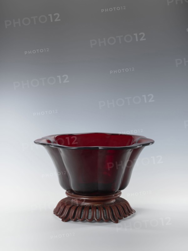 Bowl on base, bowl, Chinese art, Chinese culture, Qianlong style, Qing 1644–1911, Each segment slightly arched. The bowl's rim is rolled outward. Signed. Wooden base, carved with an openwork pattern. 1736–1795. China. Vessel. Height 12.5 cm; width 26.8 cm., A deep, translucent red glass bowl with a scalloped rim sits on a carved wooden stand, the bowl flaring outward with smooth, curved sides and a glossy surface that reflects light; the wooden stand is dark brown with an undulating, looped base and a rounded upper platform that supports the bowl, and the background is a smooth gradient of gray to light gray.