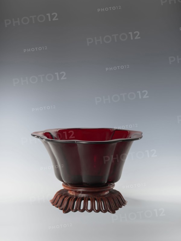 Bowl on base, bowls, Chinese art, Chinese culture, Qianlong style, Qing dynasty 1644–1911. Each segment slightly arched. The bowl's rim is rolled outward. Signed. Wooden base carved with an openwork pattern. Date: 1736–1795. Place of origin: China. Vessel. Height: 12.5 cm; width: 26.8 cm., A deep red, translucent bowl with a scalloped rim sits on a dark reddish-brown carved wooden stand with an openwork petal-like base; the bowl has smooth, curved sides and a glossy finish, and the scene shows gradients of gray in the background.