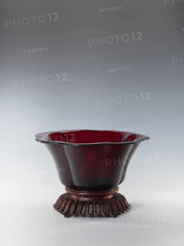 Bowl on base, bowl, Chinese art, Chinese, Qianlong style, Qing dynasty 1644–1911. Each segment slightly arched. The bowl's rim is rolled outward. Signed. Wooden base carved with an openwork pattern. Date: 1736–1795. Origin: China. Vessel. Height 12.5 cm; width 26.8 cm., A deep translucent bowl with a flared, scalloped rim sits on an ornate carved base; the bowl is a rich, dark red color with glossy highlights and the base is a darker reddish-brown wood tone with carved, looped petal-like elements.