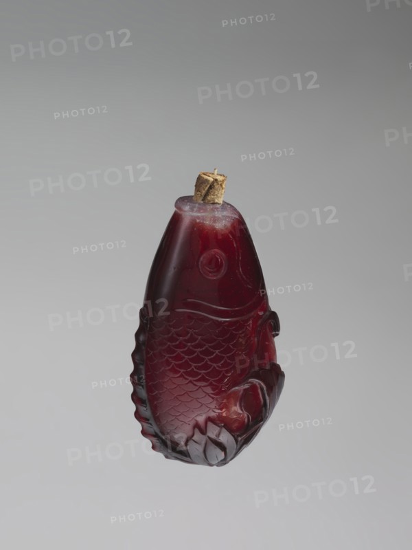 Snuffbox with spoon;, Snuffbox in the shape of a fish with a folded fin, with spoon. Porcelain or ceramic vessel; bottles motif, carps/fish imagery; Chinese art, Qing dynasty 1644–1911, 18th century, China. Height 7.0 cm, width 4.0 cm., A carved object shaped like a fish suspended against a plain background, made of a translucent deep red material with lighter red highlights and small areas of darker red shading; the fish has carved scales, a circular eye, a curved mouth area, and decorative fins, with a small brown cork or stopper set into the top.