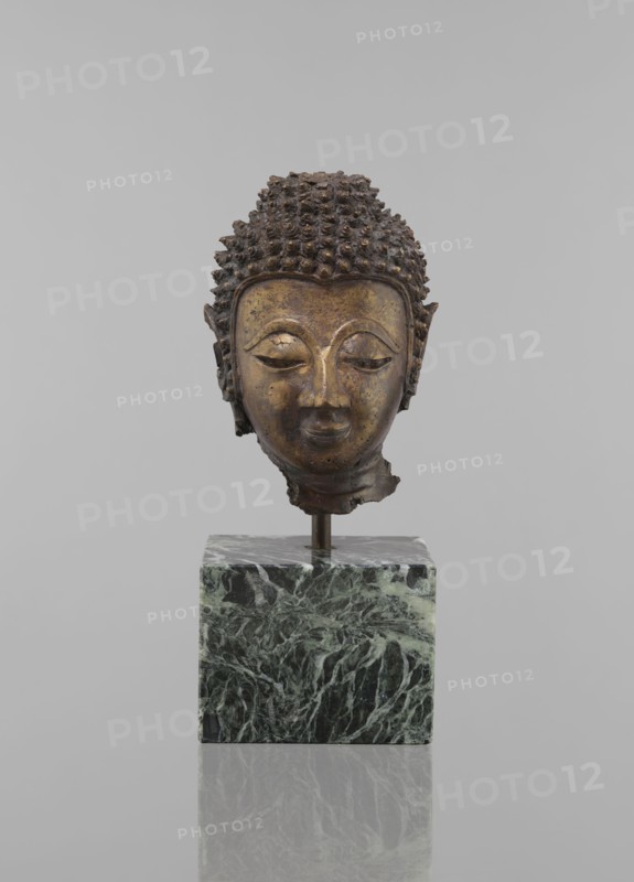 Buddha head - fragment of a sculpture, Buddhist art, Buddhism, art of Southeast Asia, Thai art, statues, fragments, ushnisha, heads, 12th–19th century, Thailand, figurine, height 11.5 cm, diameter 9.0 cm, A bronze-colored sculpted head with textured curly hair and a serene facial expression sits mounted on a metal rod above a green and black marbled cubic base; the background is a uniform light gray, and the surface beneath the base reflects the base faintly.