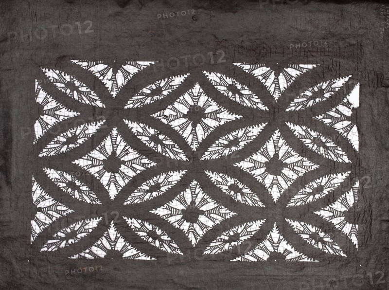 Katagami dyeing stencil with a motif of overlapping circles shippô. Ise-katagami stencil, chuban format. Pattern: geometric motif—rhombuses inscribed in ellipses with stylized floral rosettes in the rhombuses. Late 19th century Meiji period, Japan. Stencil katagami. Height 41.0 cm, width 31.5 cm., A rectangular patterned fabric with a dark gray background and white cutout areas forming a repeating geometric floral motif, featuring a grid of diamond shapes each with a central flower-like element surrounded by petal-shaped cutouts and curved leaf-like arcs; the dark gray fabric shows visible creases and texture around the patterned area, and the white cutouts reveal a contrasting bright surface beneath.