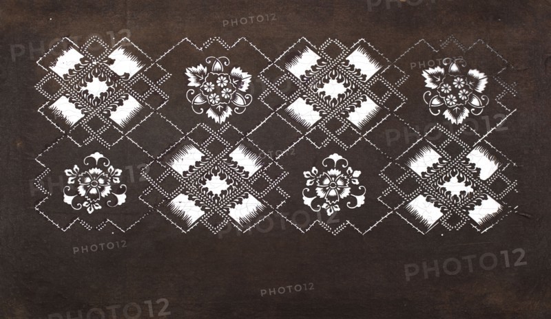 Katagami dyeing stencil with a motif of rhombuses forming crosses and plant-floral patterns hishigata jûji ni sôka moyô, Japanese, Meiji period 2nd half of the 19th century, katagami stencil, chuban format, geometric-plant patterns, flowers; stencil; height 40.7 cm, width 24.3 cm., The image shows a dark brown background with a horizontal band near the top featuring a repeating decorative pattern in white and dark brown. The pattern includes several floral-like motifs and diamond-shaped lattice elements arranged in a row, with white ornamental fills and dark brown detailing. The diamonds are connected by dashed or stitched-looking lines, and the floral motifs have leaf and petal shapes with curling flourishes. The lower portion of the image is mostly plain dark brown, while the upper patterned area contrasts with white and dark brown elements.