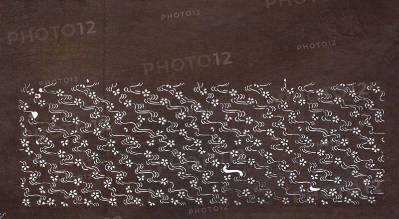 Katagami dyeing stencil with cherry blossom and flowing water motif sakura ni ryûsui, katagami stencil, chuban format; stencil of water, plant patterns, flowers, cherries sakura; 19th century Edo period, Japan; height 40.8 cm, width 22.3 cm., A dark brown textured background fills the image with a horizontal band near the bottom composed of a repeating white floral and wavy motif; the band contains a group of small stylized flowers and flowing curved lines in white that form a dense patterned area against the brown, with occasional isolated white shapes punctuating the pattern.