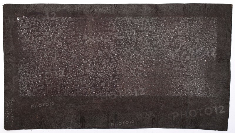 Katagami dyeing stencil with bamboo motif take, Japanese, Edo period 19th century, katagami stencil chuban format, plant pattern of bamboos, stencil, height 43.1 cm, width 24.0 cm., A rectangular sheet with a dark brown surface and a slightly lighter brown central panel filled with a repeating dotted floral pattern, bordered by a wider dark brown frame with a textured, crinkled appearance; a few small whitish spots interrupt the patterned area, and the outer edge rests on a pale off-white background.