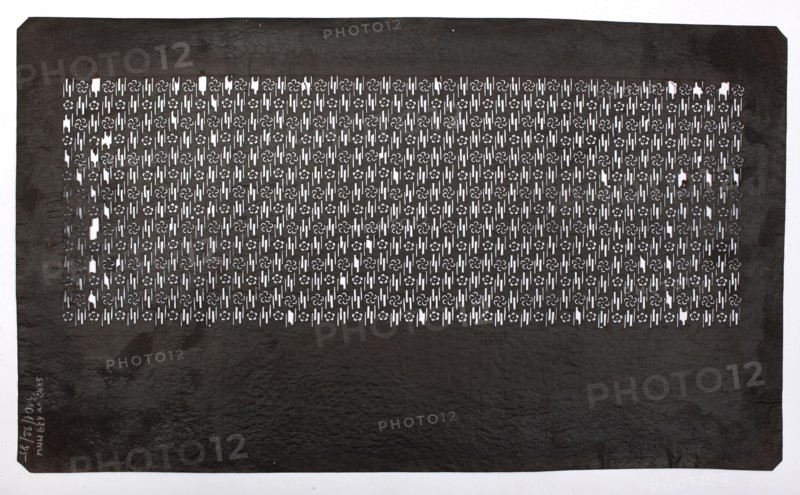Katagami dyeing stencil with plum blossom and stylized mist motif ume no hana ni kasumi moyô, katagami stencil, chuban format; geometric and plant patterns, flowers, plum trees; stencil; Meiji period, 2nd half of the 19th century Japan; height 40.3 cm, width 23.7 cm., A large rectangular sheet with a dark brown to almost black surface fills the image, showing a patterned central panel of cut-out shapes that reveal a white background beneath; the cut-outs form a repeating decorative motif across a horizontal band near the top, leaving a broad plain darker area below. The patterned band displays a grid of small floral-like shapes and narrow vertical slits, creating a high-contrast interplay of white and dark. The sheet has slightly irregular edges with small corner bevels and subtle surface texture variations, including faint lighter spots and scuffs. Overall the visible colors are dark brown to black for the sheet and white showing through the pattern.