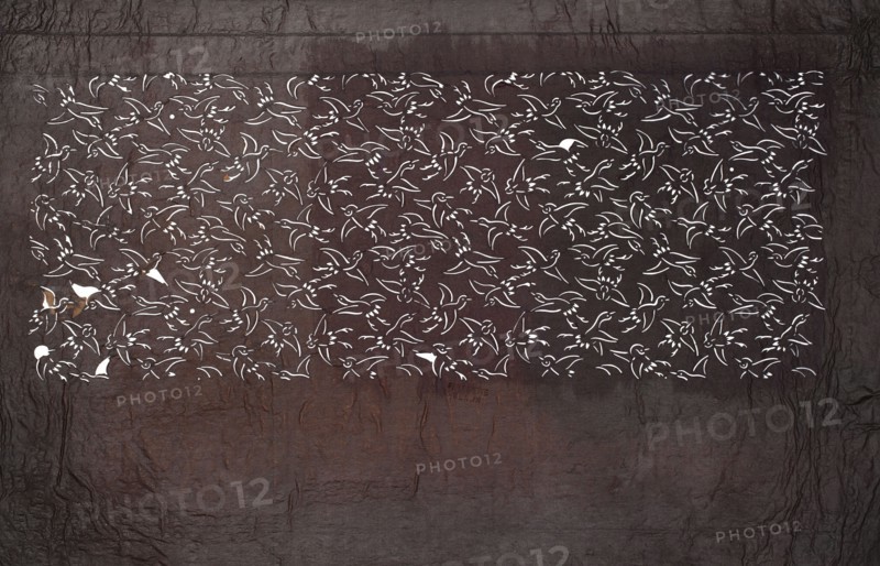 Katagami dyeing stencil; pattern of densely arranged birds in flight, ise-katagami stencil, Meiji period 1868–1912, late 19th–early 20th century, Japan; stencil; height 39.7 cm, width 24.7 cm; Japanese art, stencils., A rectangular section filled with a repeating bird motif cut out in white sits against a textured dark brown and gray background; the birds are stylized with simple outlines and wing shapes, appearing in a continuous pattern across the rectangle, and there are a few small areas where the white cutouts reveal underlying rust-colored patches; the surrounding surface shows crinkled, leathery texture in shades of deep brown, charcoal gray, and muted reddish-brown.