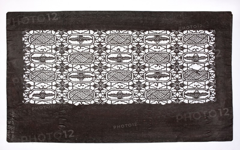 Katagami dyeing stencil; geometric-floral pattern stylized after the traditional Chinese brocade pattern syokko; maker, Japanese; Meiji period 1868–1912; ise-katagami stencil with brocade ornaments; 19th–20th century; stencil; height 40.3 cm, width 23.6 cm., A rectangular sheet with a dark border surrounds an inner panel of intricate cutwork forming a repeating geometric and floral pattern; the border is a deep brownish-black and shows subtle texture and creases, while the inner cutwork is dark against a bright white background, creating high contrast. The cutwork panel contains a grid-like arrangement of oval and octagonal shapes alternating with stylized floral motifs and small decorative curlicues, all rendered in the same dark color as the border, with areas of white visible through the openwork. The overall composition is flat and centered within a clean white surrounding, and the visible colors are dark brownish-black and white.