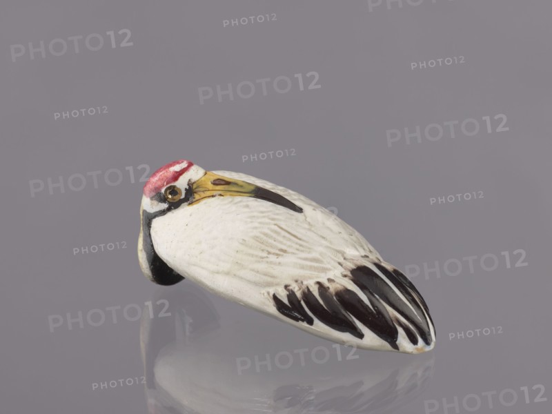 Vase, unknown ceramicist; Meiji period 1868–1912, Japan; ceramics; vessel vase; subject: cranes iconography; dimensions: height 9 cm, diameter 3.5 cm; 19th/20th century., A small ceramic bird figure resting on a reflective surface, showing a rounded body with carved feather details; its main body color is white with dark brown and black markings on the tail feathers and wing tips, a yellowish beak with a darker tip and a small dark eye area, and a red patch on the top of its head, all set against a muted grayish-lavender background.