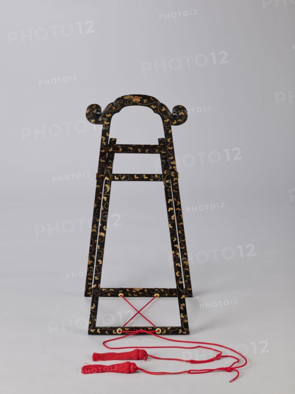 Mirror stand kagami-kake, wooden stand covered with black lacquer; painted with gold with plant ornaments, mirrors, Edo period 1st half of the 19th century, Japan, height 66 cm, A decorative black wooden stand with curved top elements and scroll-like ends, covered in a gold floral and vine pattern, standing on a light gray surface against a pale gray background, with a few red cords and tassels resting in front of it.