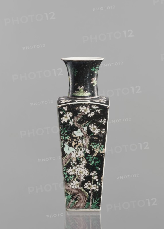 Pair of vases, Chinese, 18th century, colored porcelain famille noire, decorated with birds, plums and floral motifs; vessel, height 22.5 cm., A tall, narrow ceramic vase with a flared rim and a square shoulder, decorated on a dark background with floral and bird motifs; the surface shows branches with blossoms in white and pale pink, leaves in shades of green, and pale green birds perched among the flowers, with brownish tree trunks and subtle green foliage at the base; the rim and interior appear white, and the overall background is black with some lighter worn areas, set against a neutral gray backdrop.