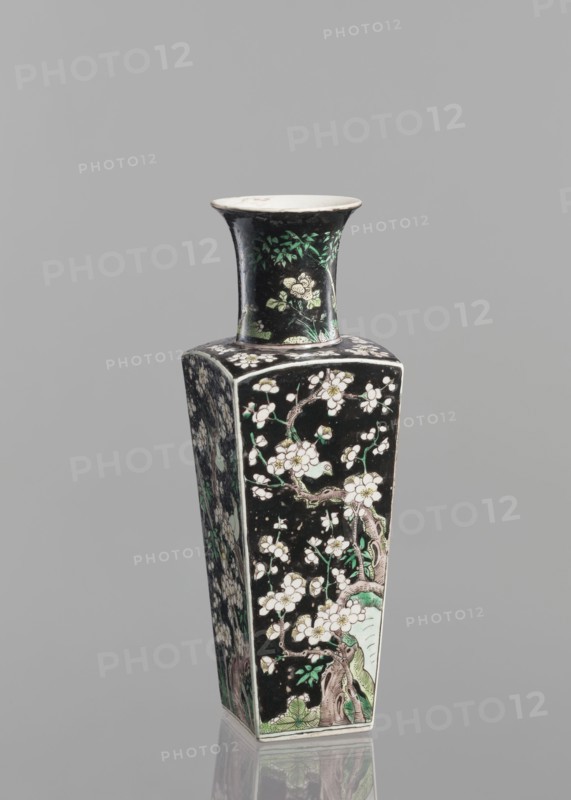 Pair of vases; Chinese; 18th century; colored porcelain famille noire; decorated with birds, plums, and floral motifs; vessel; height 22.5 cm; ., A tall, square-based ceramic vase with a flared rim, shown against a plain gray background. The vase has a predominantly black surface decorated with floral and tree branch motifs in white, green, and pale pink, with brownish outlines and some lighter green foliage near the base and neck. The neck features a cluster of green leaves and pale flowers, while the body panels display blossoming branches and leaves in white, green, and pale pink set against the black ground. The rim interior and the very top edge are a pale off-white color. The vase sits on a reflective surface that shows a faint mirror image of its lower section.