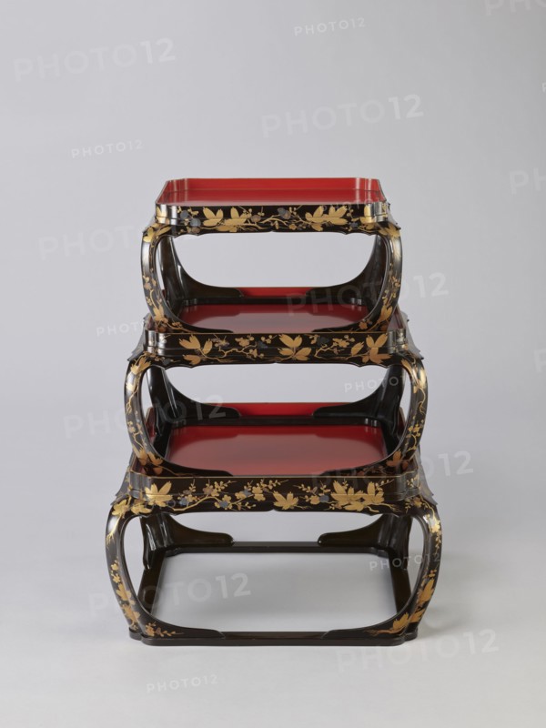 Set of three trays on legs kakeban. Japanese, Edo period early 19th century. Low tables, identically decorated but of different dimensions allowing them to be stacked pyramidally, one on top of the other. Each table has an almost square top framed with high slats, supported by four curved legs connected at the bottom by four slats forming a square base. The top and the inner side of the slats framing it are coated with vermilion-coloured lacquer; the remaining surfaces are covered with shiny black roiro-nuri lacquer and decorated with a golden ornament of pine branches and plum blossoms made using the sprinkling technique hiramaki-e; decoration slightly raised above the background surface. Subject motifs: plum trees, flowers, trees, pine trees, twigs, winter. Piece of furniture., A three-tiered stand with curved rectangular frames appears centered against a pale gray background, the stand showing a glossy black finish with decorative gold floral and leaf patterns along the edges and su...