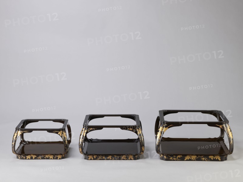 Set of three trays on legs kakeban. Japanese, Edo period, early 19th century. Low tables, identically decorated but of different dimensions allowing them to be stacked pyramidally. Each has an almost square top framed with high slats, supported by four curved legs connected at the bottom by four slats forming a square base. The top and the inner side of the slats framing it are coated with vermilion-coloured lacquer; the remaining surface is covered with shiny black roiro-nuri lacquer and decorated with a golden ornament of pine branches and plum blossoms made using the sprinkling technique hiramaki-e; decoration slightly raised above the background surface. Subject: plum trees, flowers, trees, pine trees, twigs, winter. Piece of furniture., Three low, rectangular stands sit side by side against a neutral light gray background, each with a glossy dark brown or black lacquer finish and curved open sides. The stands form a tiered shape with a flat top frame and a lower shelf, and they fe...