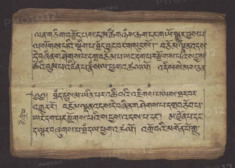Buddhist manuscript — Penitential Text Tib. sDig bshags gser gyi zhes bya ba bzhugs+o, unknown creator / scribe-copyist, Tibetan Buddhist art, Himalayan culture. Book in Tibetan pecha style, manuscript in student's script. Leaf numbering 2–22 verso, first recto, last two unnumbered. Handwritten notes folio 24 recto and verso. 19th century, Tibet. Manuscript; width 24.0 cm, height 8.1 cm., An open aged book page with a worn cream-colored paper showing dark brown edges and layered, slightly darker brown pages at top and bottom; the page contains a group of black handwritten script arranged in two horizontal blocks separated by a faint horizontal crease and a few small dark spots; thin vertical margin lines frame the text on both sides and a few ink marks appear near the left margin; the background around the book is a deep dark brown.