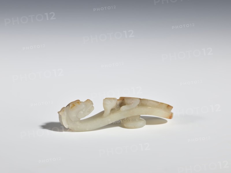 Belt clasp, lizards zool., belt clasps, Chinese, Chinese art, Qing dynasty 1644–1911, dragon iconography, 19th century, China; belt buckle; height 6.5 cm, width 1.8 cm., A small carved object in pale off-white and creamy tones with areas of light brown and tan patina rests on a smooth white surface; it has a curved elongated form with rounded protrusions and a bulbous base, subtle translucent areas, and soft shadowing beneath, set against a gradient background that transitions from light gray to darker gray at the top.