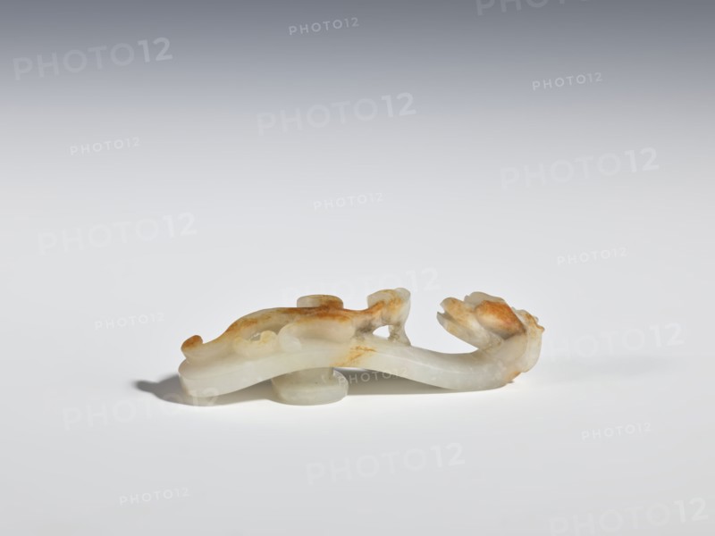 Belt clasp, purchase provenance; materials and form: belt clasp/belt buckle depicting lizards and dragon iconography; culture: Chinese, Qing dynasty 1644–1911, 19th century, China; dimensions: height 6.5 cm, width 1.8 cm., A small carved object rests on a flat surface, shaped with flowing, curved forms and rounded protrusions. The object appears smooth and translucent with areas of warmer, rusty brown and cream tones blending into pale white and light gray. Its silhouette suggests an elongated body with a thicker rounded section toward one end and a curled, sculpted detail at the other. The surface shows subtle variations in opacity and mottled patches of color, and a soft shadow falls beneath it on the white-gray background.