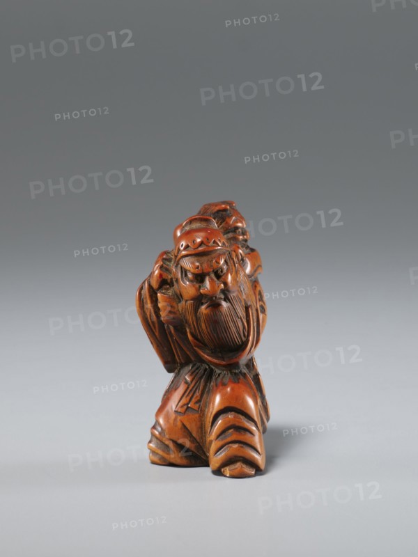 Netsuke in the shape of Shoki and an oni; maker, Japanese, Edo period 1st half of the 19th century; carved netsuke sculpture; subject: Shoki mythological figure and a demon; materials: carved netsuke; dimensions: height 4.5 cm, width 3.0 cm., A small carved figurine with a polished brown and reddish-brown surface depicting a bearded man wearing a rounded hat and flowing garments, shown from the front with a pronounced facial expression and detailed carved hair and beard, standing on a base formed by the folds of his clothing; the background is a smooth gradient of gray tones.