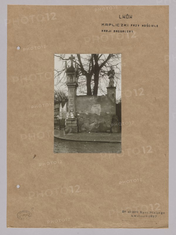 Chapel at the Church of St. Mary Magdalene in Lviv, with sculpture of John of Nepomuk on the wall of the former Dominican church; photograph by Józef Kosciesza Jaworski fl. ca. 1911–ca. 1929; negative dated 1911–1918, print dated 1911/1918–ca. 1922; photographic print glued to brown cardboard print: 15.7 × 10.6 cm; support: 35.2 × 25.6 cm; photography, nature photography., A sepia-toned photograph mounted on a brown paper sheet shows an outdoor scene with a weathered wall and a gate structure; the central image features a stone pillar topped with a cross on one side and a statue on a pedestal on the other, with bare tree branches rising behind them. The ground in front appears to be a paved street or sidewalk with a curved curb, and the wall surface shows patches of peeling plaster and uneven texture. A few posters or paper remnants are visible on a section near the left pillar. The sheet around the photograph is a warm brown color with a slightly rough texture and small punched holes ...