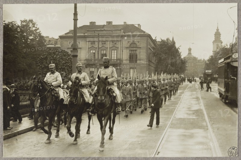 1. Warsaw. German cavalry in the street, surrounded by children, August 5, 1915; 2. Warsaw. German regiment and people on Krakowskie Przedmiescie, near Zamkowy Square, August 5, 1915; 3. German troops marching along Krakowskie Przedmiescie, past the Tyszkiewicz-Potocki Palace. Photographer: Alfred Kühlewindt 1870–1945. Three photographs glued together on a single cardboard; 5 August 1915. Subjects and details: occupation, World War I 1914–1918, Warsaw Masovian Voivodeship, trams, Krakowskie Przedmiescie street, German army, horses, palaces, soldiers, children, Warsaw inhabitants, Sigismund's Column, Staszic Palace, Church of the Holy Cross, St. John the Baptist. Photography; Poland; Warsaw., A sepia-toned street scene shows a mounted group of men in uniform riding horses at the front of a marching formation, with a larger group of uniformed soldiers walking behind them holding long rifles. A few civilians stand or walk alongside the street, and a tram or streetcar is visible on the rig...