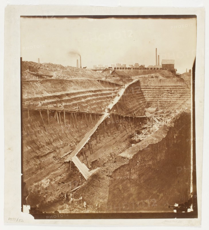 Open-pit hard coal mine Cieszkowski in Dabrowa Górnicza - view from the east, Fajans, Maksymilian 1825-1890 photographer, Light-sensitive, coal mines, Art of photography, Dabrowa Górnicza Silesian Voivodeship, miners, industrial landscapes, Zaglebie Dabrowskie Silesian Voivodeship, Print glued to cardboard with its entire surface, 1864 references to the taking of the photograph: Tygodnik Ilustrowany, 1864, no. 256 of August 20, p. 307; Gazeta Warszawska, 1864, no. 192 of August 23, p. 1, Dabrowa Górnicza place of origin, nature photography, photograph, photographic print: height 42.5 cm, width 37.7 cm; cardboard: height 47.2 cm, width 42.9 cm, A sepia-toned photograph showing a large excavated pit with stepped, terraced walls and exposed soil layers, a long wooden plank ramp supported by vertical posts zigzagging down into the pit, a few small figures of people working along the ramp and at the bottom, scattered wooden beams and debris in the lower area, a raised flat area at the top w...
