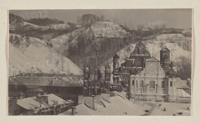 Churches and mountains; photograph from the album Vilnius, volume 1; Jan Bulhak 1876–1950; Vilnius, 1912; scenes: Bernardine Church Vilnius, St. Anne's Church Vilnius, St. Michael's Church Vilnius, panoramas; photographic print: 9.5 × 16.3 cm; album page: 31.0 × 23.5 cm., The image shows a snowy scene with a group of rooftops and ornate church towers in the foreground and a hilly, wooded landscape behind them. Visible colors include shades of gray, off-white, beige, and muted brown. The foreground contains several pitched roofs covered in snow, chimneys, and scaffolding near a light-colored building with decorative architectural details. Clustered behind and beside that building are darker, more ornate spires and domes with layered shapes and pointed tops. In the background a series of pale, snow-covered hills rise, punctuated by darker tree lines and bare trees on the slopes, creating a contrast between the lighter hills and the darker wooded ridges. The whole scene is framed by a pal...