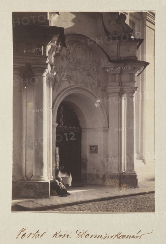 Dominican Church portal; photograph, Bulhak, Jan 1876–1950, Vilnius Lithuania, baroque style, Dominican Church of the Holy Spirit Vilnius, 1912; photographic print: height 15.5 cm, width 11.1 cm; album page: height 31.0 cm, width 23.5 cm, The image shows a stone building entrance with decorative architectural details, including an arched doorway framed by columns and ornate relief above the arch; a dark interior is visible through the doorway and a person wearing a dark garment is seated or crouched near the entrance; the scene includes a paved street in front and strong light creating highlights and shadows on the columns and facade; visible colors include shades of beige, cream, brown, gray, and black.