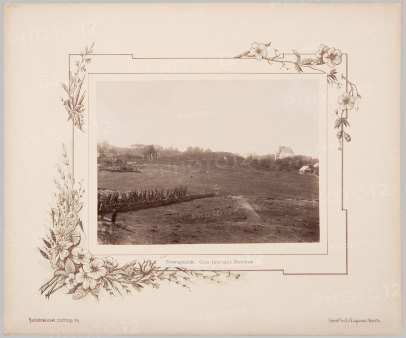 Nowogródek. Mindaugas Mountain Mound. Album card with photographic print glued onto lithographed cardboard. Photography from nature, print height 16.3 cm, width 22.5 cm; album card height 33.0 cm, width 39.8 cm. 1894. Subjects/notes: landscapes; mounds; Mindaugas Mountain Nowogródek; Nowogródek Belarus; Lithuania; Minsk; Adam Mickiewicz; makers/authors: Józef Mikolaj Boretti photographer, Teofil Eugeniusz Boretti compiler/author., A sepia-toned photograph shows a wide open grassy landscape with gently rolling hills, a group of taller plants or crops forming a line across part of the midground, and a few small buildings or houses visible on the distant hillside; near the foreground a solitary figure stands beside the planted row. The photo is mounted on a pale beige mat decorated with an ornate floral border featuring drawn flowers and leaves in brown and cream tones at the corners and along the sides. The overall visible colors are sepia, beige, brown, and cream.