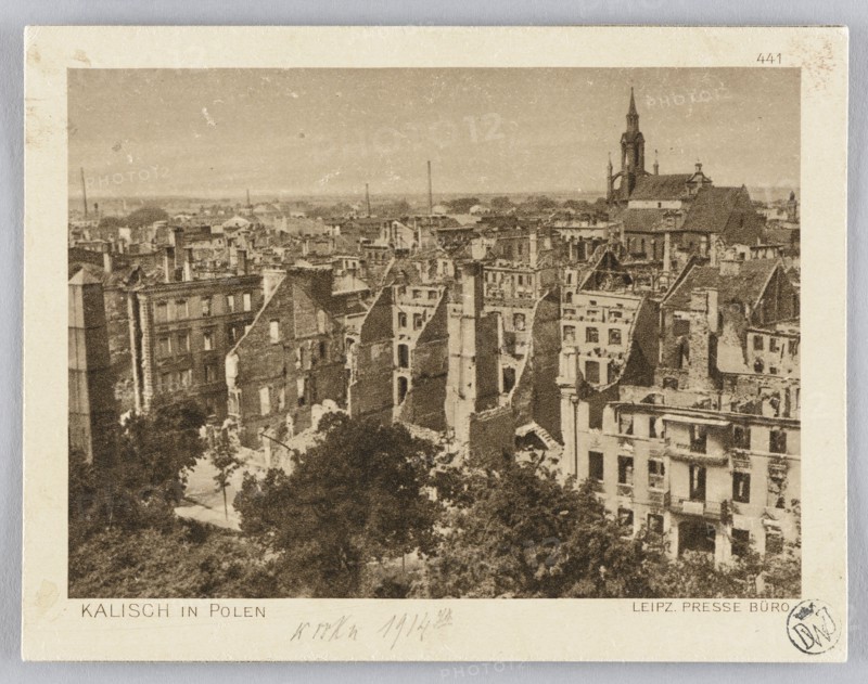 Panorama of Kalisz after its destruction in 1914; panorama city view showing war damage and churches. Leipziger Presse-Büro press office original author; JF Lehmanns Verlag Munich publisher. Postcard glued across the entire surface onto a cardboard backing; postcard, 1914. Germany place of origin; Munich place of publication. Dimensions: height 9.3 cm, width 12.0 cm., A sepia-toned photograph shows a dense urban scene of damaged and partially ruined multi-story buildings with many exposed walls, broken roofs, and missing sections, creating jagged rooflines and interior views; a tall church with a steeple is visible rising above the cluster of buildings in the background; there is a group of trees and foliage in the foreground partially obscuring lower parts of the buildings; the overall palette includes shades of brown, beige, tan, and darker brown tones across the structures and vegetation.