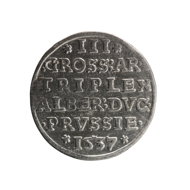 Trojak, Vogelwaider Vogelwaid, Eustachius; publisher Decius, Jost Ludwik fl. 1528–1535 co-creator; mintmaster. Obverse: male bust in armour in profile turned right; short hair, beard with sideburns; rim inscription *ALBERDG MAR BRAN DVX PRVS with continuous border and floral motifs between words. Reverse: continuous border with six-line inscription *III*/.GROSS:AR:/.TRIPLEX./ALBER:DVC./.PRVSSIE./*I537*; dated 1537. Ducal Prussia 1525–1701, Königsberg mint. Coin: diameter 22.1 mm, weight 2.63 g, axis 50°., A round metallic object with a worn silver-gray surface fills the frame against a white background; the object shows raised, blocky shapes arranged in horizontal rows and small decorative marks near the edges, with surface scratches and varied patina giving darker and lighter gray areas across its face.