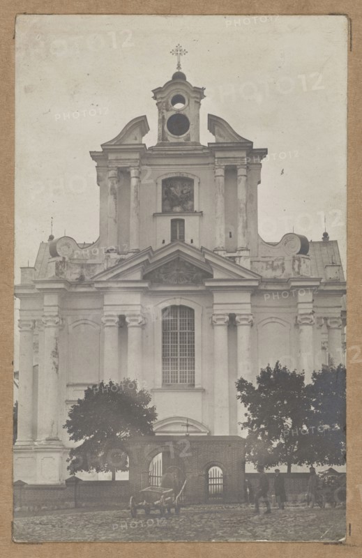 Sienno Sianno, Belarus. Dominican Church of St. Trinity — general view towards the façade, architecture, gates, churches, sacred architecture, lost monuments, general views. Photographer unknown. ca. 1910–1913. Print glued to brown cardboard. Photography from nature; photographic print: 13.8 × 8.7 cm; cardboard: 35.3 × 25.5 cm., A large classical stone building with columns and decorative curved roof elements dominates the image, shown in a muted grayscale. The facade includes a tall central window with a grid of panes, an ornate triangular pediment above it, and a round opening near the top beneath a small tower topped by a cross. There is visible weathering and texture on the building surfaces. In front of the building is a low brick wall with an arched opening flanked by metal gates, and a group of dark-leaved trees on either side. On the cobblestone street in front are a few people standing and walking, and a couple of horse-drawn carts with visible wheels and harnesses. The overal...