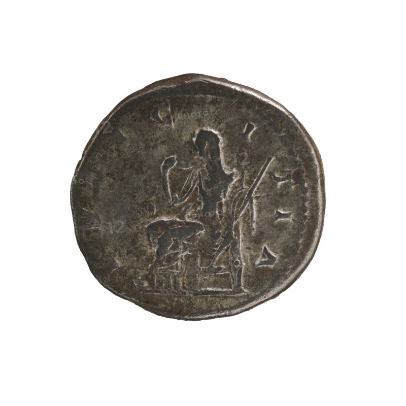 Denarius, Elagabalus 204–222; Roman emperor 218–222. Obv.: IVLIA MAESA AVG — Draped bust of Julia Maesa facing right, pearl border. Rev.: PVDICITIA — Pudicitia with a veil on her head sitting on a throne facing left, lifting the hem of her veil with her right hand and holding a scepter in her left, pearl border. 218–223, Roman Empire; Rome mint. Coin; diameter 19.5 mm, weight 3.50 g., A round metallic coin with a dark brown and gray patina showing wear and uneven surface texture; the coin features a raised, worn relief of a seated human figure facing left with an arm raised and draped clothing, and a smaller raised form near the figure that suggests an object or small stool; there are faint raised marks and irregularities around the edge of the coin and variations in the metal color including areas of darker brown, dull gray, and subtle greenish tones.